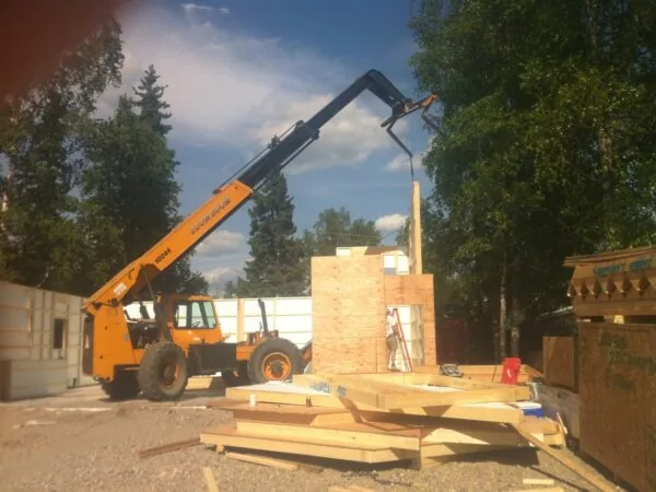 Telescopic handler lifting wooden materials at a construction site with brick structures and trees.