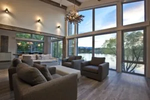 Modern living room with floor-to-ceiling windows overlooking a lake, featuring gray upholstered furniture and a chandelier.