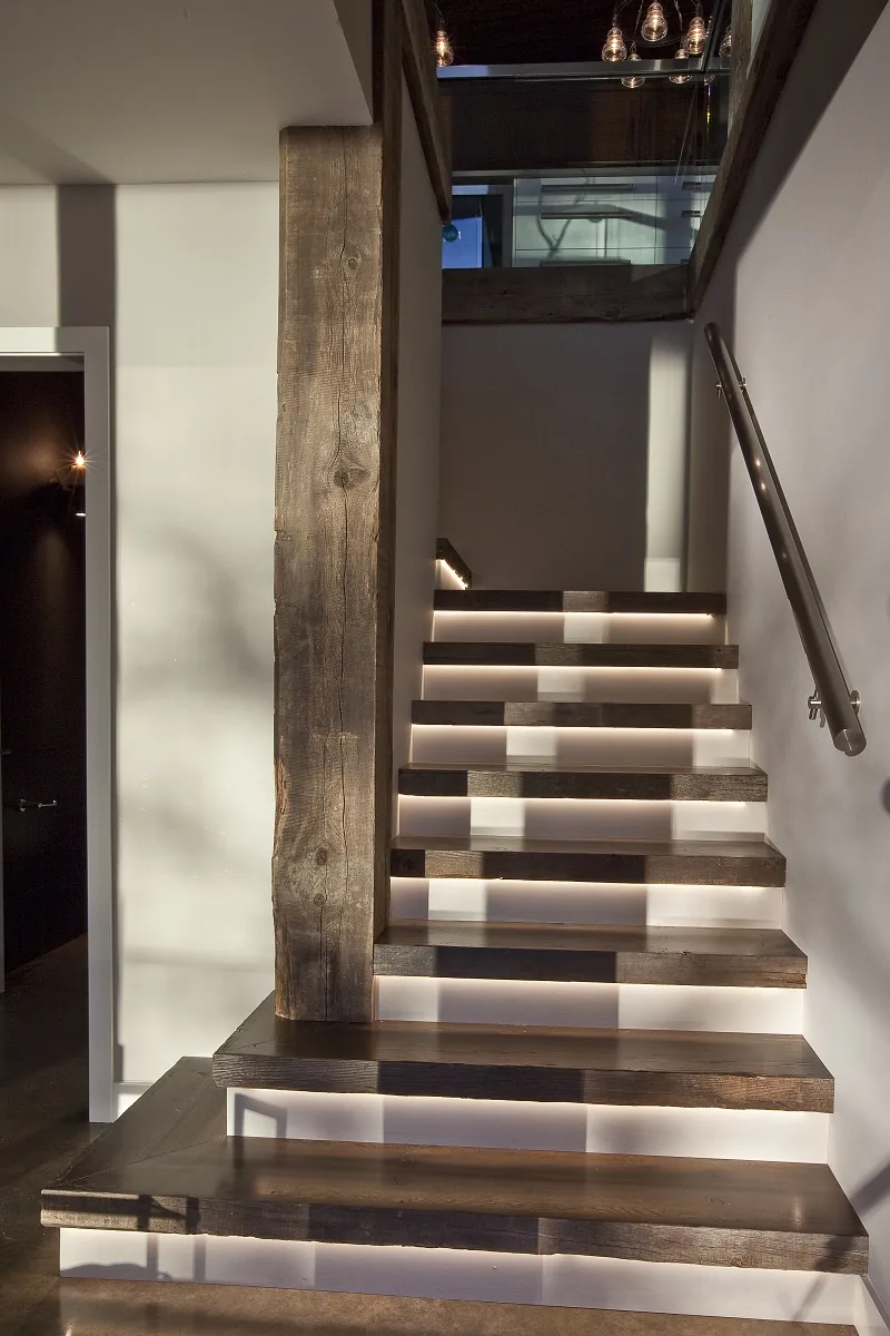 Modern staircase with dark wooden steps and recessed lighting beneath each tread, featuring a weathered wood support column and metal handrail.