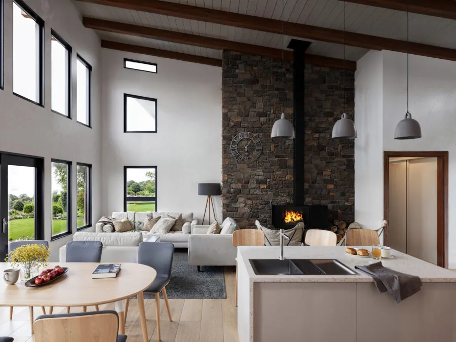 Modern open-plan living space with brick fireplace, kitchen island, and large windows overlooking green landscape. (2 of 4)