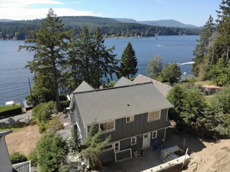 Waterfront home with gray roof nestled among pine trees overlooking a blue lake with mountains in the distance.
