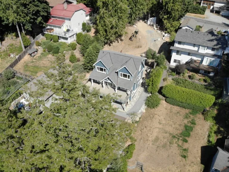 Aerial view of a two-story blue house with a driveway surrounded by trees and neighboring properties.