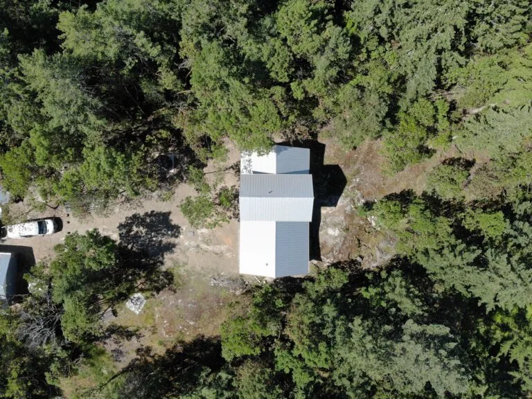 Aerial view of a modern white and gray house surrounded by dense green forest with a gravel driveway.