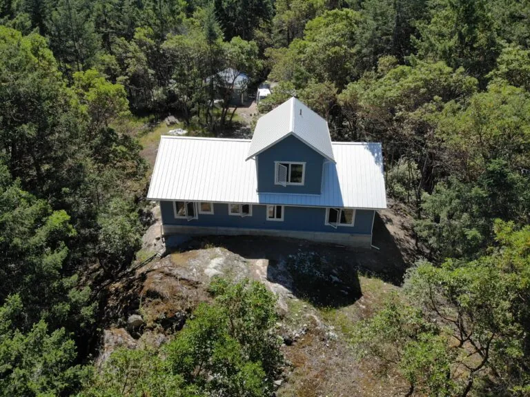 A modern house with white metal roof and dark blue siding sits atop a large boulder surrounded by dense forest.