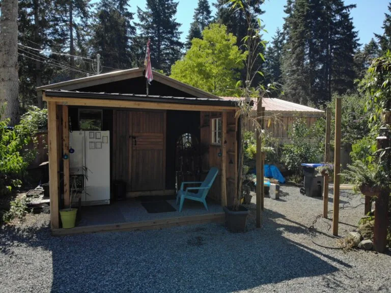Rustic wooden cabin with open front door, white refrigerator, blue Adirondack chair, and gravel patio surrounded by tall evergreen trees.