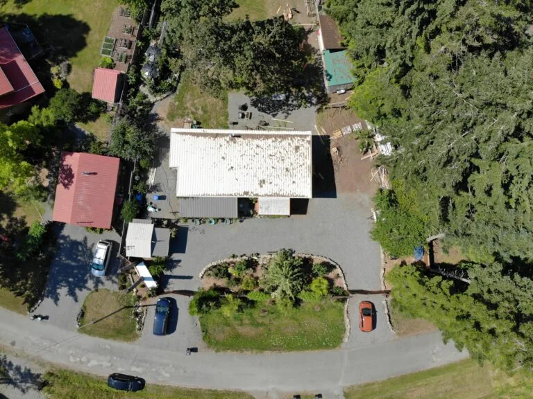 Aerial view of a residential property with a large white warehouse building surrounded by forest and parked vehicles on a curved driveway.
