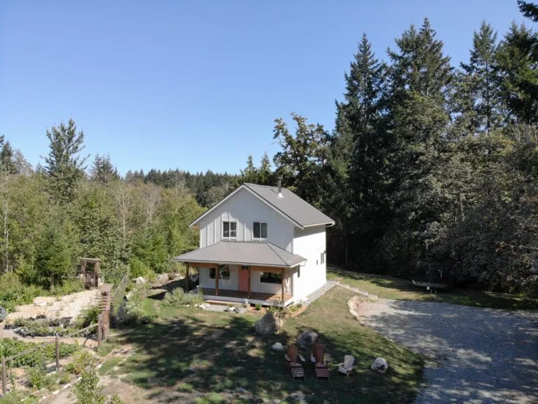 Modern white farmhouse surrounded by evergreen forest with gravel driveway and front porch.