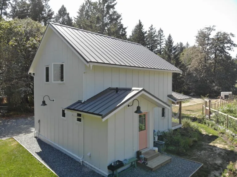 Modern farmhouse with metal roof, white siding, and front entrance surrounded by trees and fenced garden area.