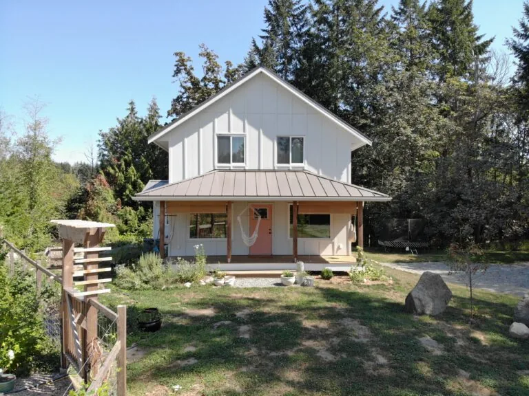 Modern farmhouse with white exterior, metal roof, and covered porch surrounded by tall evergreen trees and natural landscape.