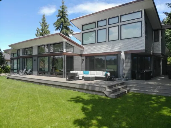 Modern two-story house with extensive glass walls and a composite deck overlooking a manicured lawn.