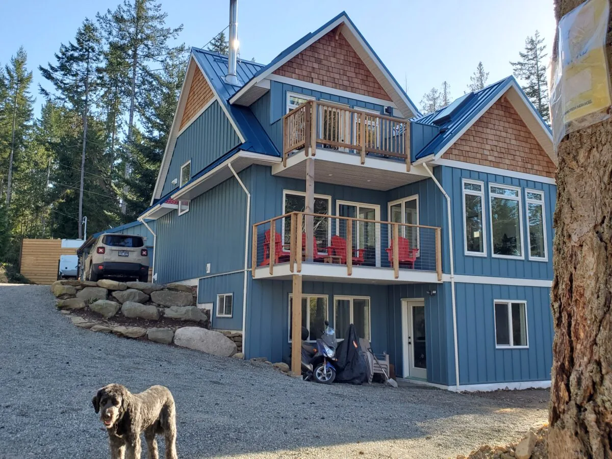 A blue modern house with cedar shake accents and multi-level decks sits among pine trees, with a dog in the foreground and a car parked nearby.