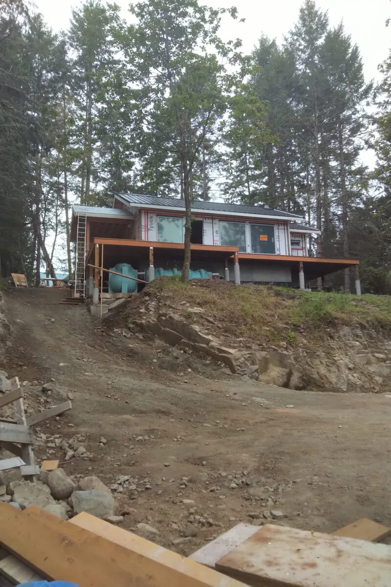 A modern cabin with orange railings sits on stilts on a hillside surrounded by tall evergreen trees.