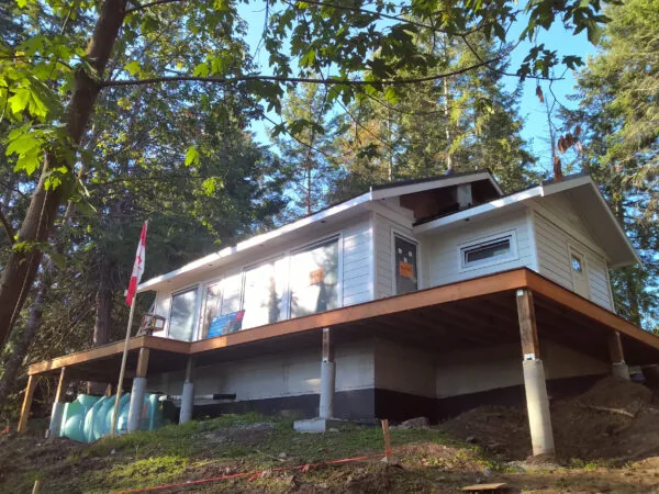 Modern elevated house nestled among tall trees with wooden deck and white siding.