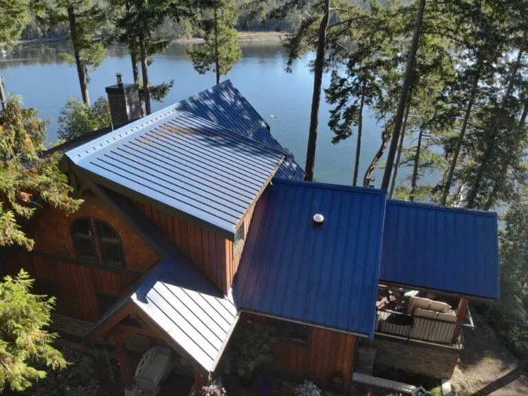 Aerial view of wooden cabin with blue metal roof nestled among trees overlooking a calm lake.