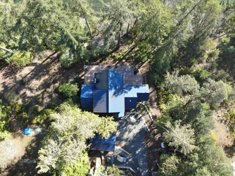 Aerial view of a modern house with blue and white metal roofing surrounded by dense forest trees.