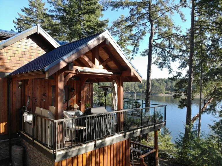 Modern wooden lakehouse with covered porch overlooking calm water surrounded by tall pine trees.