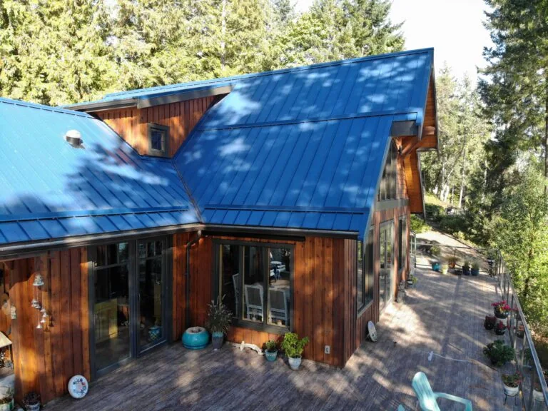 Modern cabin with dark blue metal roof and wooden siding surrounded by evergreen trees and a wooden deck.